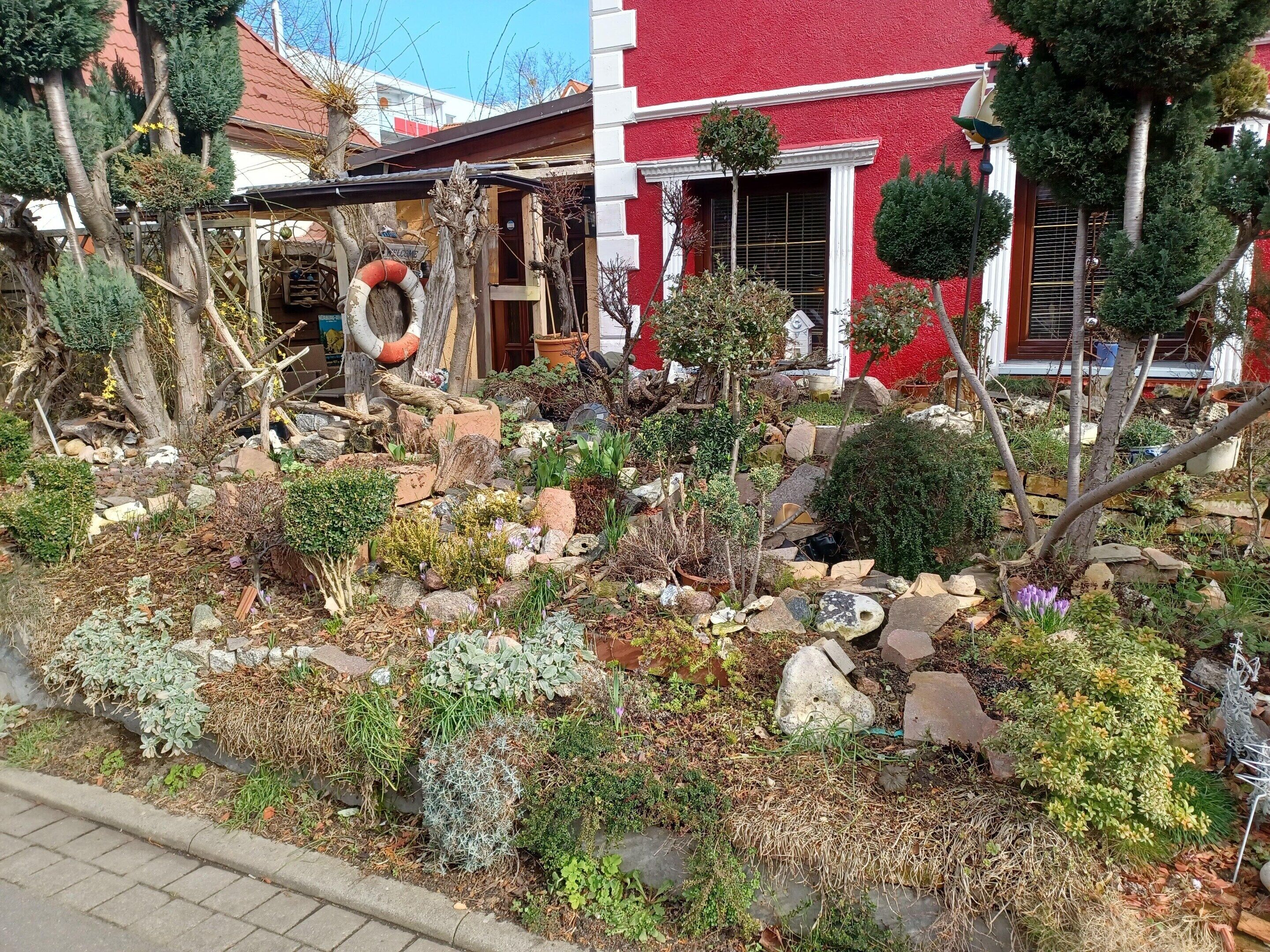 Ein Garten mit verschiedenen Pflanzen und Steinen, ein rot gefärbtes Gebäude mit weißer Verzierung sowie eine Holzhütte mit einem Schild.
