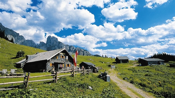 Un pueblo montañoso rural con casas tradicionales de madera, cielo despejado y una bandera.