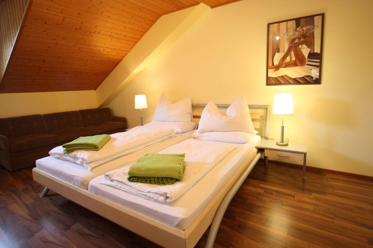 Una camera da letto con soffitto in legno, un letto con lenzuola bianche, un asciugamano verde, un divano marrone e un quadro appeso al muro.