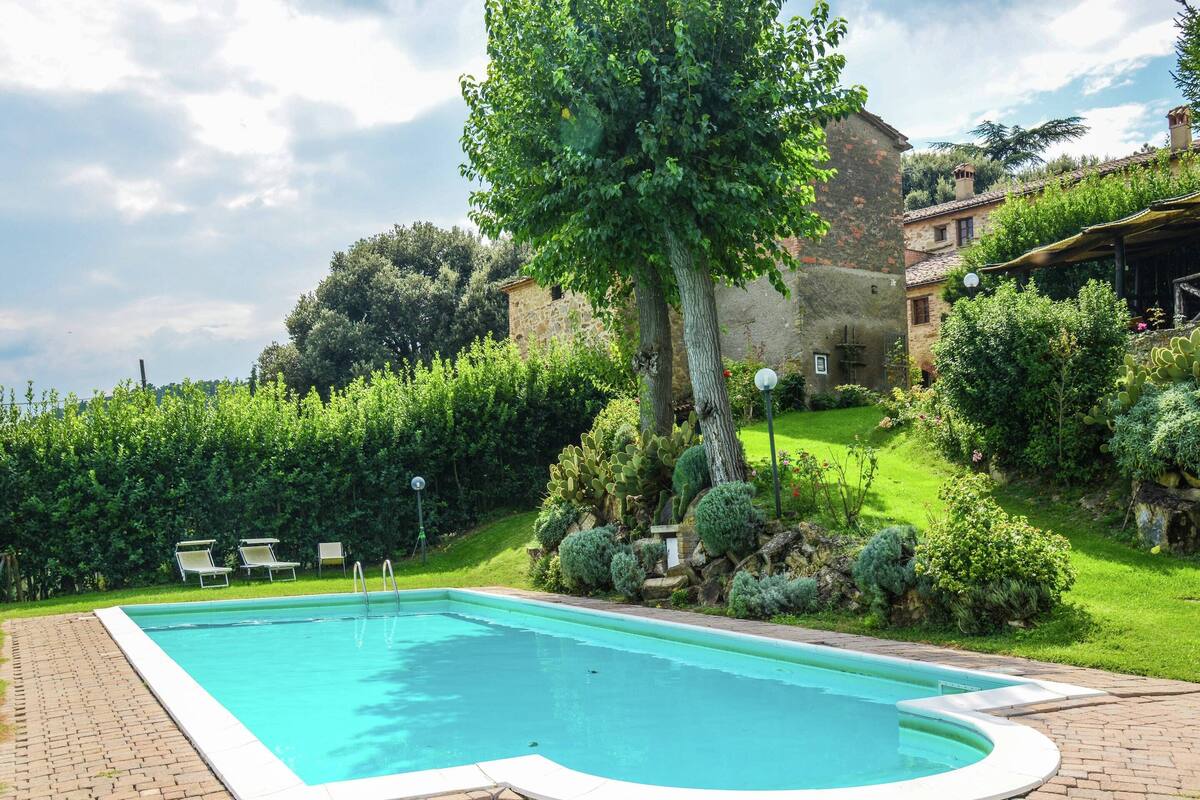 A rectangular swimming pool with a white border, surrounded by a brick patio. There are lounge chairs and a well-maintained garden with various plants and a large tree.