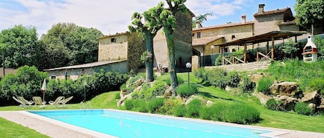 A swimming pool with a paved deck, surrounded by lush greenery and stone buildings.
