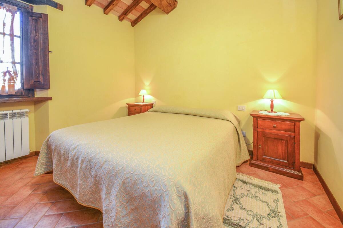 A bedroom with a bed, two bedside tables with lamps, a wooden ceiling, a radiator, and a window with curtains.