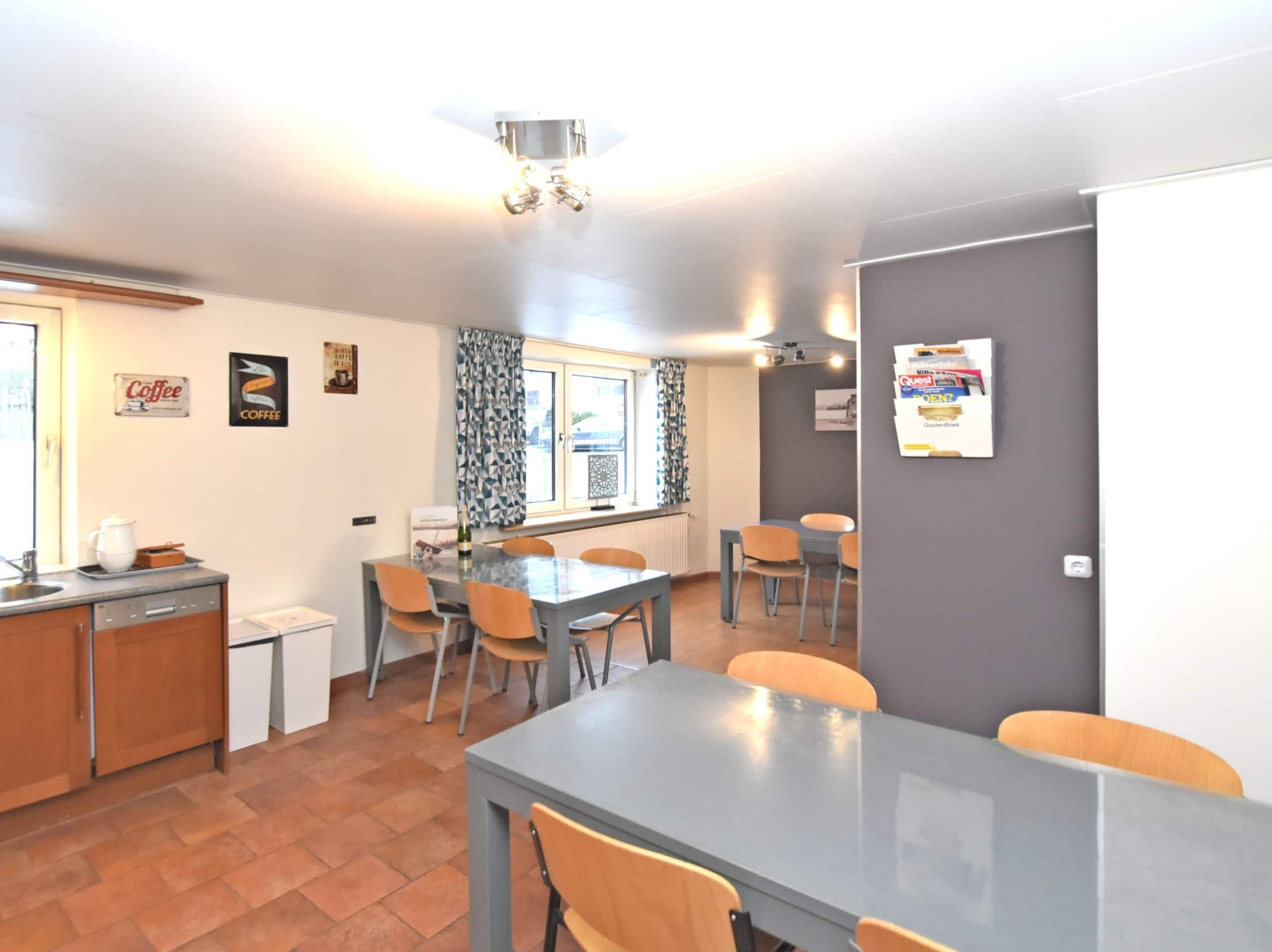 A modern kitchen with a dining area, featuring a large table and chairs, a coffee station, and a window with patterned curtains.