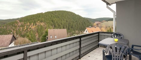 A balcony with a view of a forested hill, a table with chairs, and a white tiled floor.