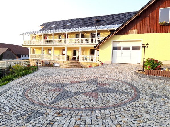 A building with a balcony, a garage, and a cobblestone driveway. - Ferienwohnungen mit Teich in Lichtenhain (Sebnitz)