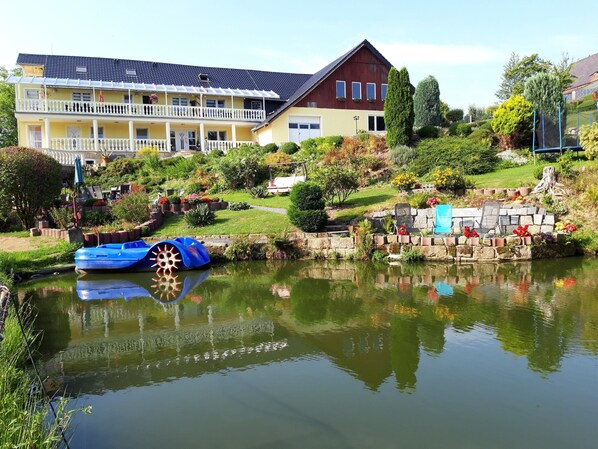 Ein Wohngebäude mit Balkon, einem Teich mit blauem Boot und einem Garten mit verschiedenen Pflanzen und Blumen.