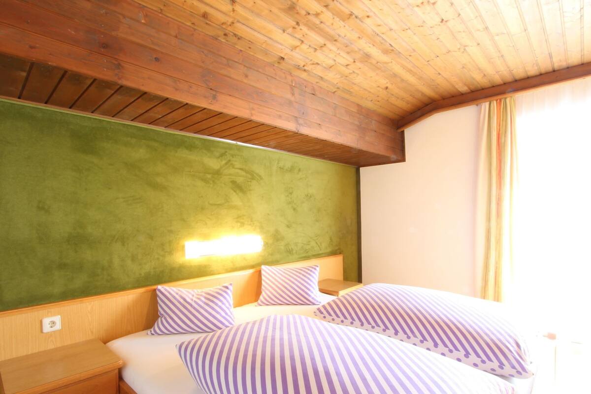 A hotel room with a wooden ceiling, a green accent wall, a bed with striped bedding, a nightstand, and a wall-mounted light.