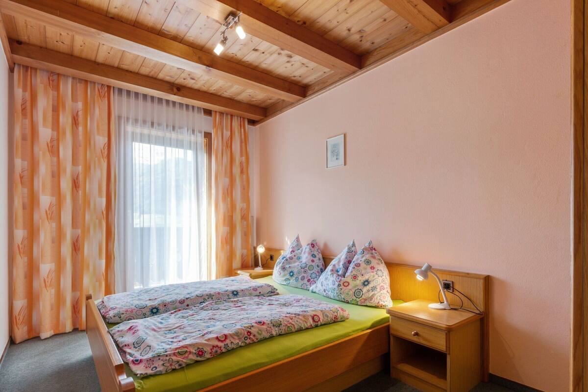 A bedroom with a wooden bed, floral bedding, a nightstand with a lamp, and a window with sheer curtains.