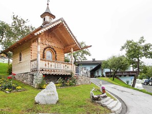 Eine hölzerne Kapelle mit einem Glockenturm, umgeben von einem Blumengarten und einem Steinmonument.