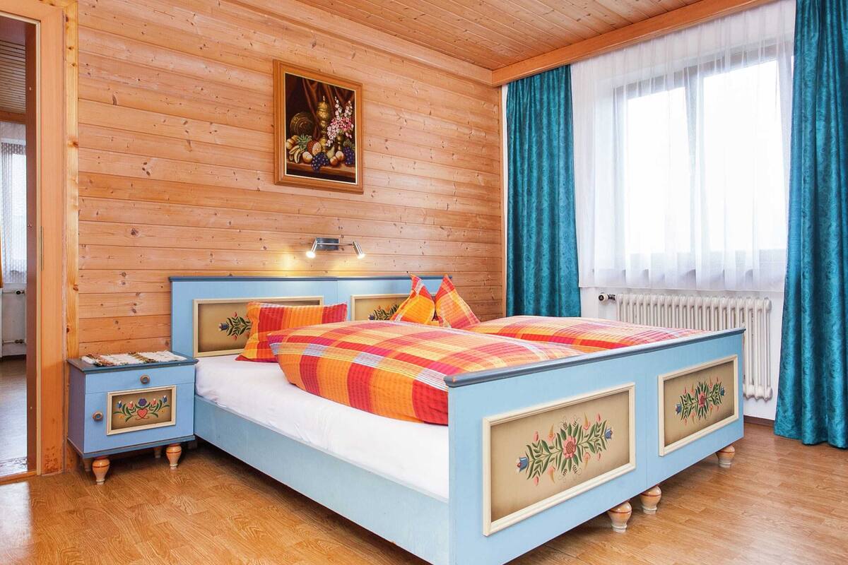 A bedroom with a wooden ceiling and walls, a bed with a colorful quilt, a nightstand with a decorative drawer, and a window with curtains.