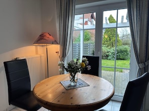 A round wooden table with a vase of flowers, two black chairs, a lamp, and a view of a garden through a glass door.