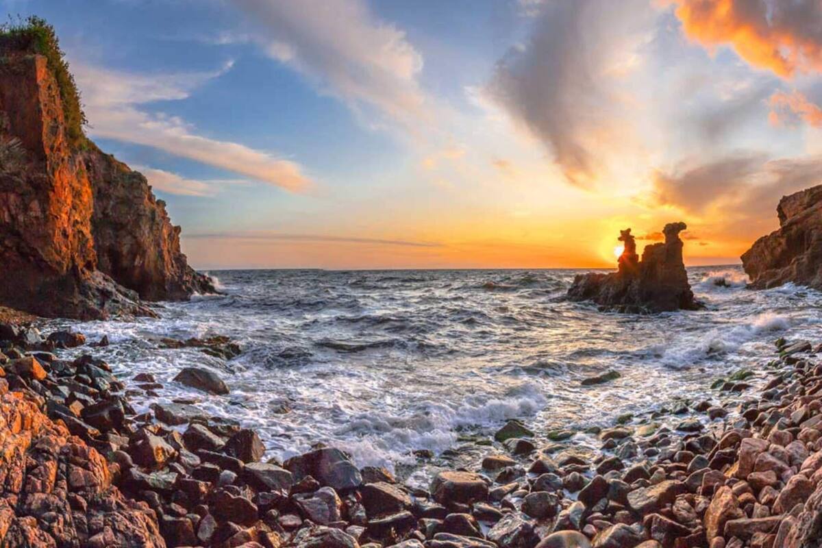 A rocky coastline with waves crashing against the shore and a sunset in the background.