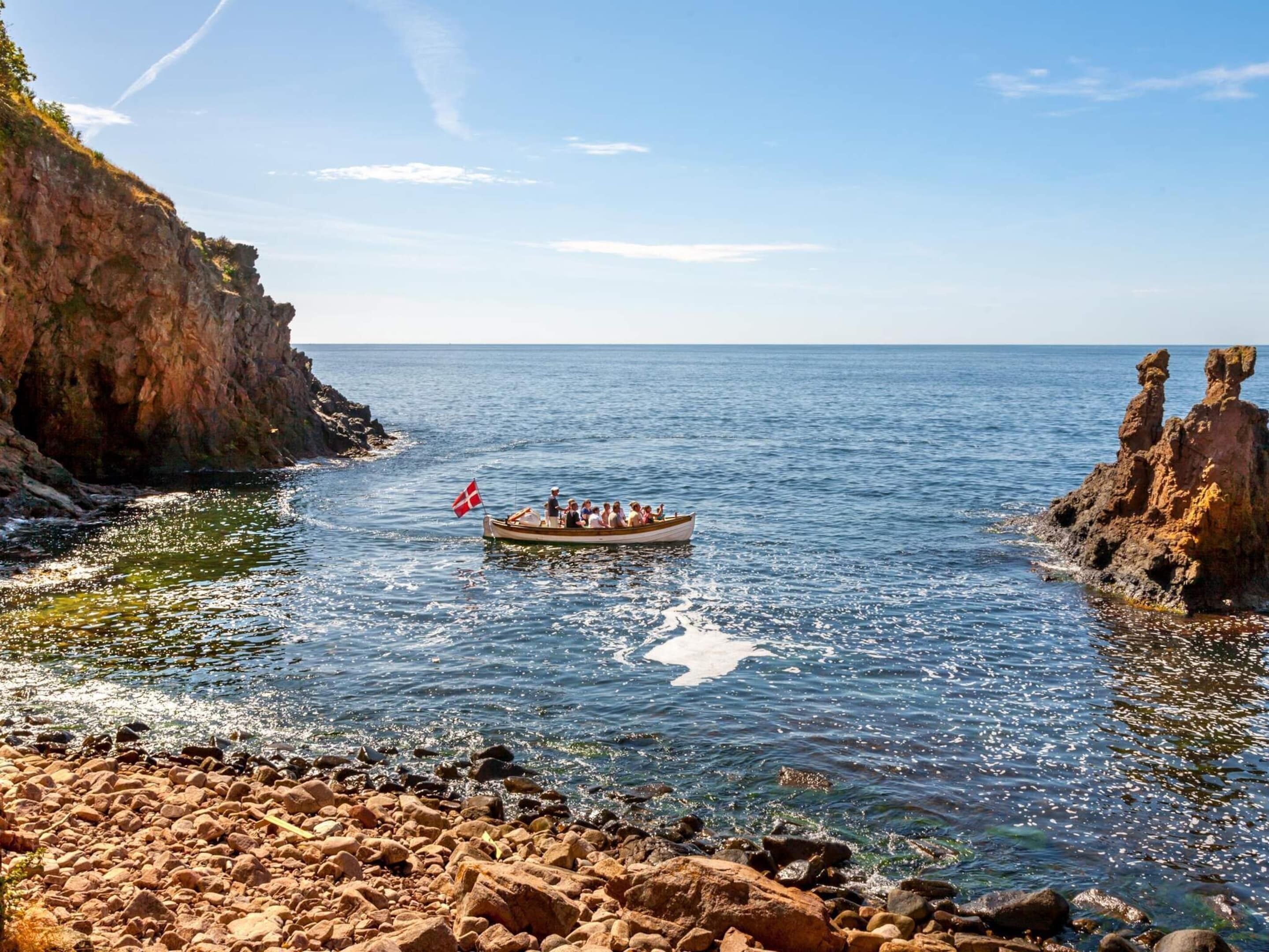 A rocky shoreline with a boat carrying passengers and a flag, surrounded by calm sea and clear skies.
