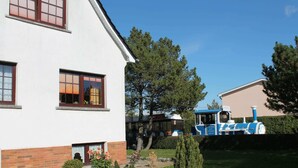 A two-story house with a brick base and white upper walls, a small garden with various shrubs, and a blue and white train in the background. - Charming Apartment in Kuhlungsborn With Parking (Kuehlungsborn)