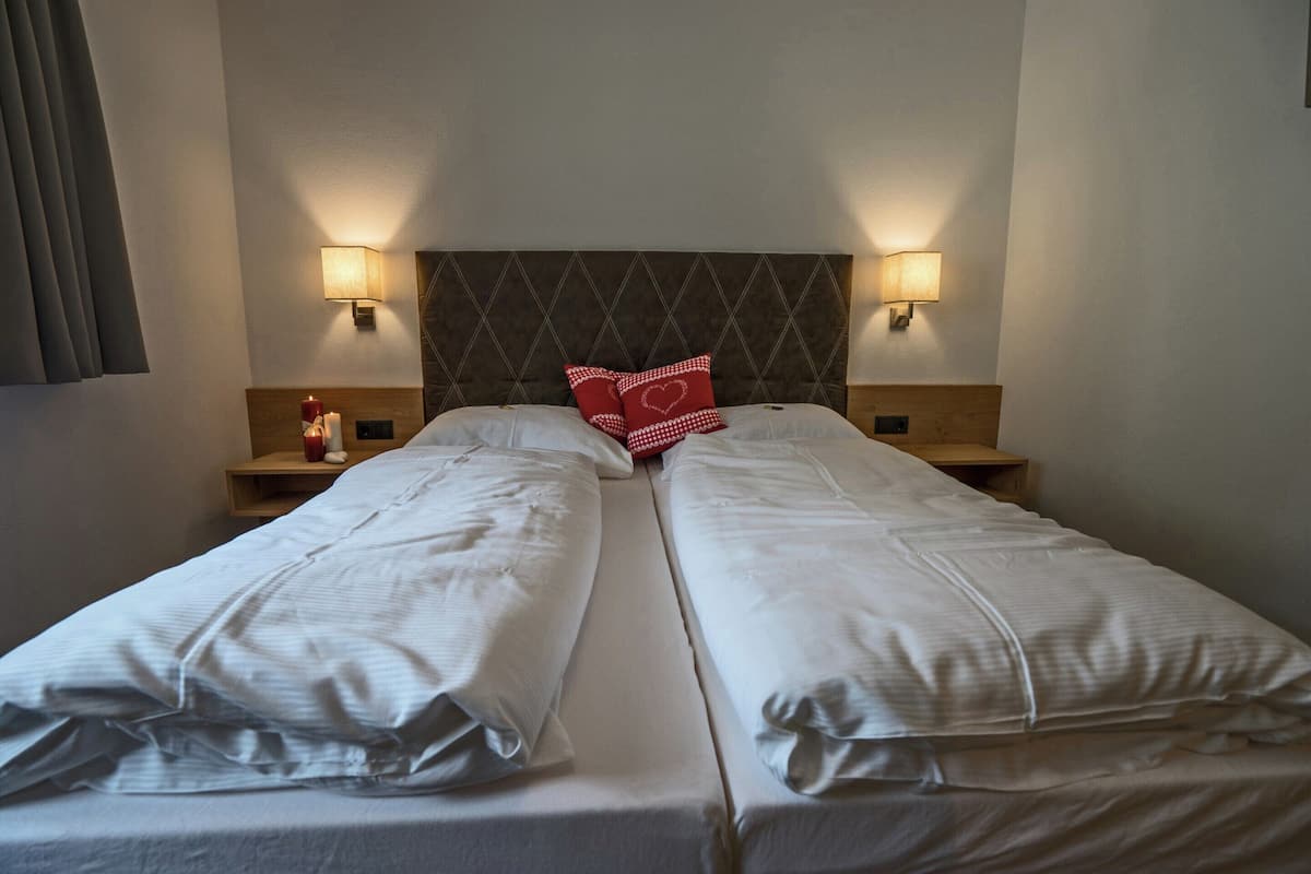 A double bed with white linens and a red heart pillow, two bedside lamps, and a wooden nightstand with a bottle and a candle.