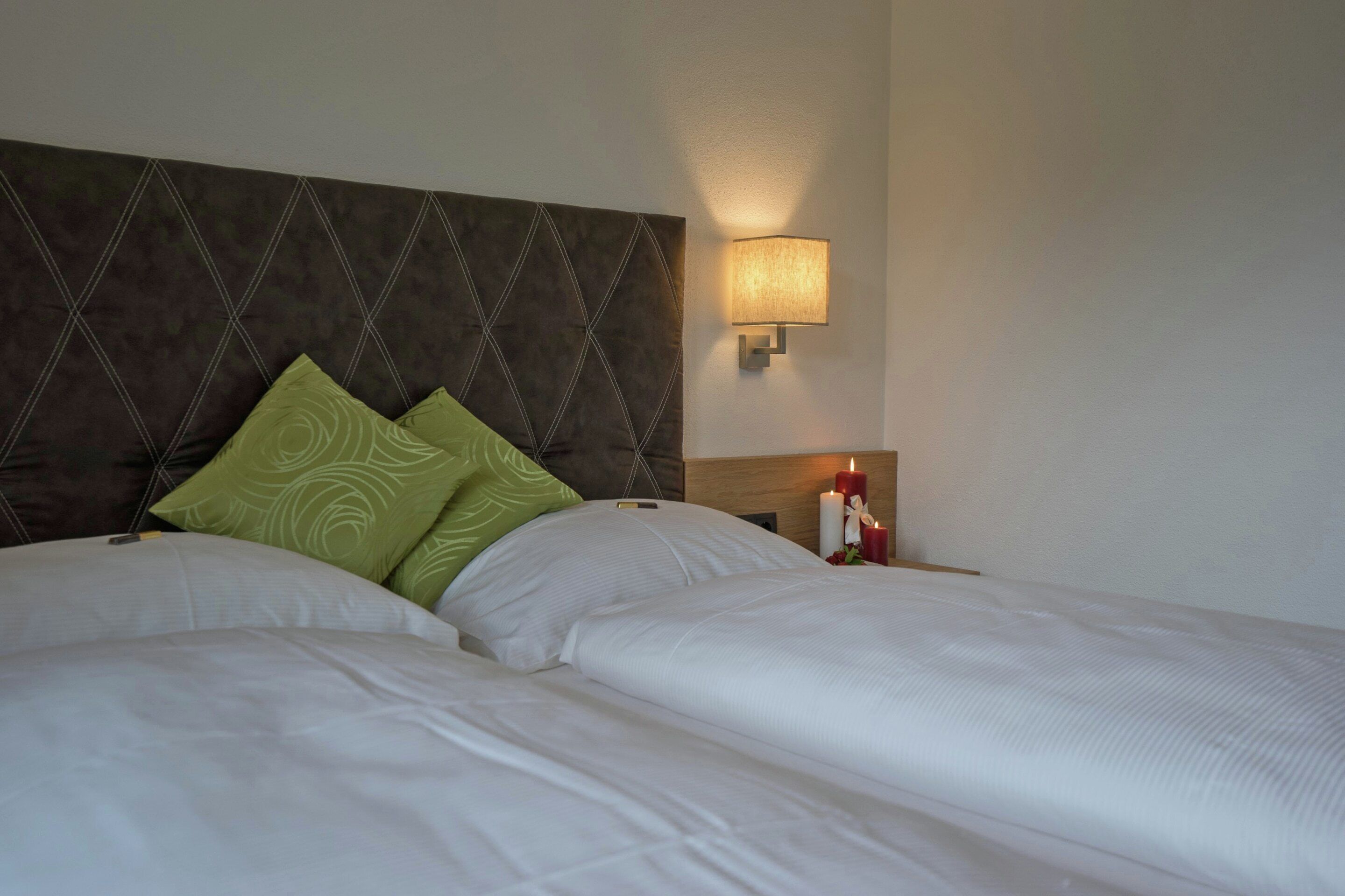 Habitación de hotel con dos camas, cabecero con diseño de rombos, lámpara de pared y mesita de noche con una vela.