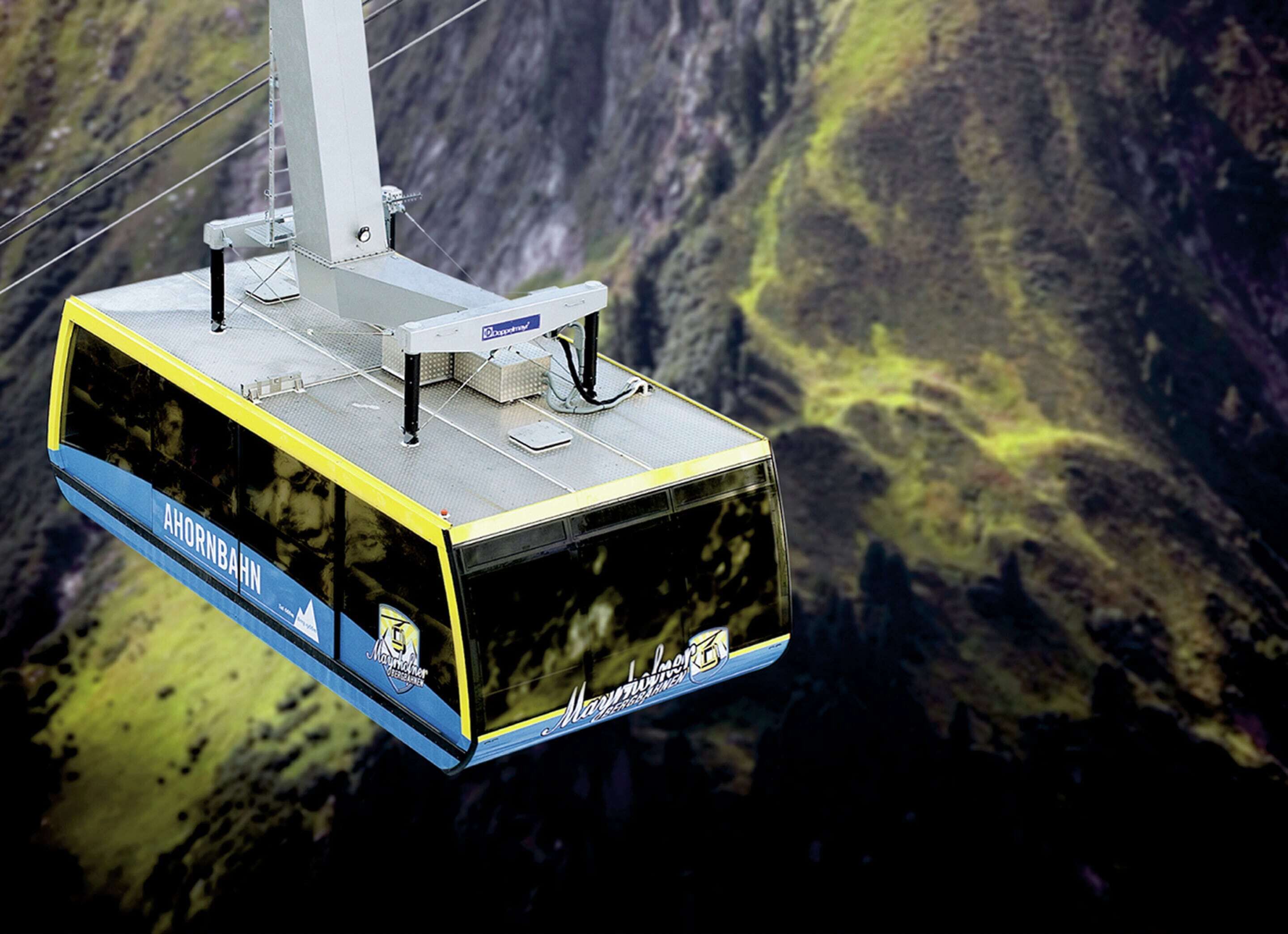 A cable car with "AHORNBAHN" and "Mayrischboden" visible, suspended above a mountainous landscape.