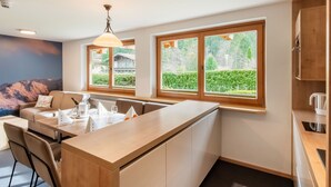 A modern kitchen with a central island, large windows with a view of greenery, and a dining area with a sofa and chairs. - Lovely Apartment With Sauna, Ski Storage, Pool, Terrace (Wald im Pinzgau)