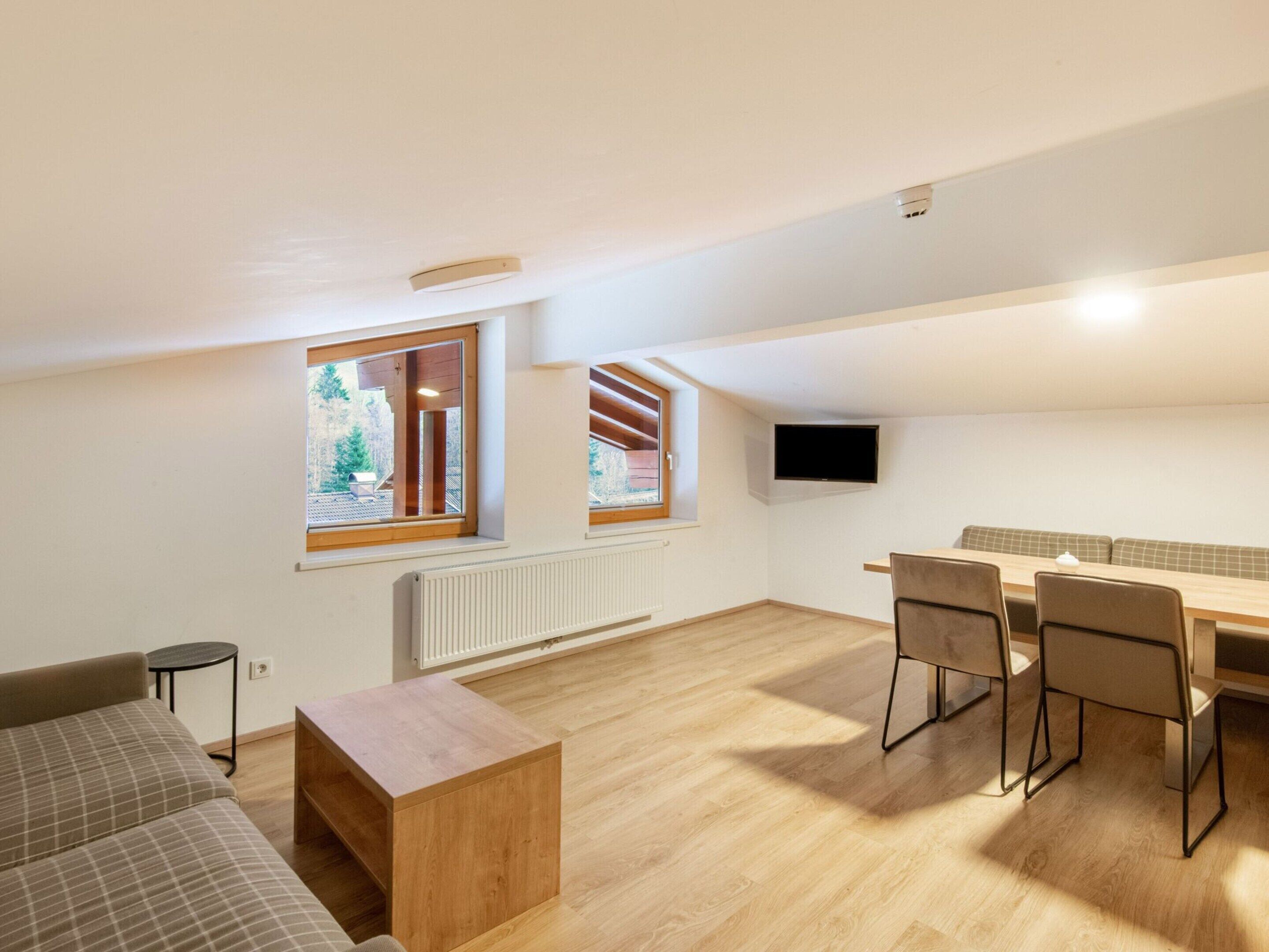 A modern, well-lit apartment with a dining area, a sofa, a TV mounted on the wall, and large windows offering a view of the outdoors.