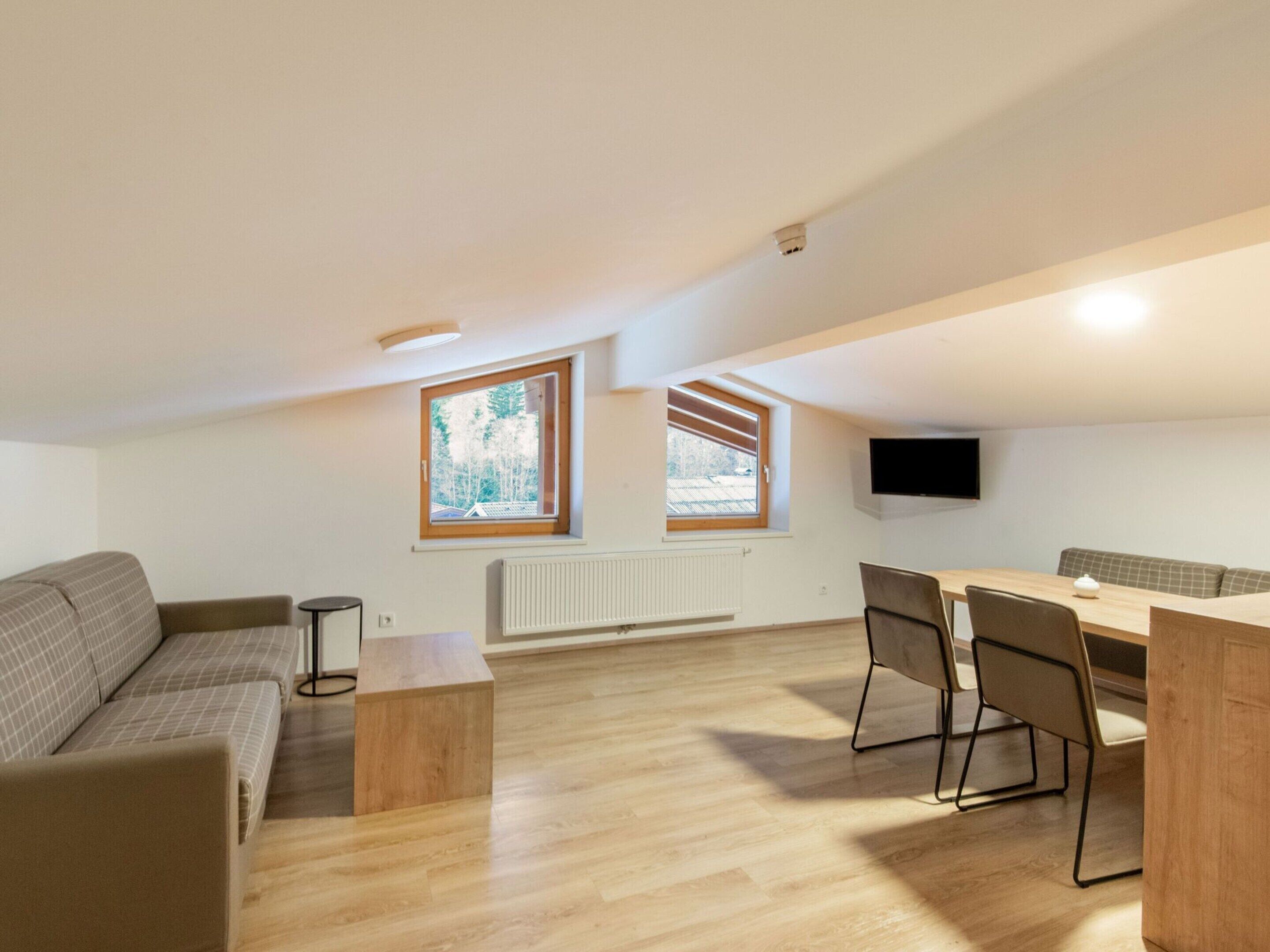 A modern, open-plan living and dining area with a wooden floor, a sofa, a dining table with chairs, and two windows with blinds.