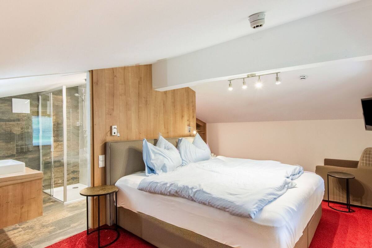 A modern hotel room with a large bed, a glass shower enclosure, a small round table, and a red carpet.