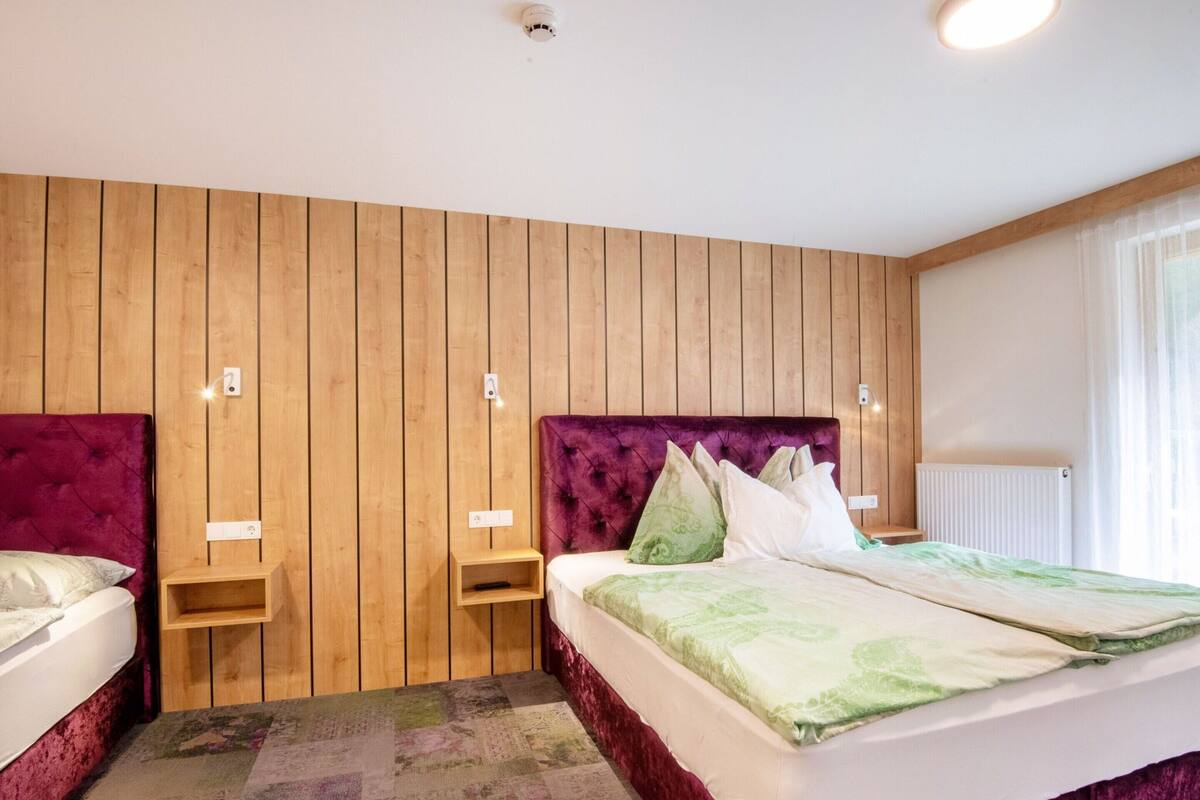 Une chambre d’hôtel avec un mur lambrissé en bois, un grand lit doté d’une tête de lit violette, deux tables de chevet et une fenêtre avec des rideaux.