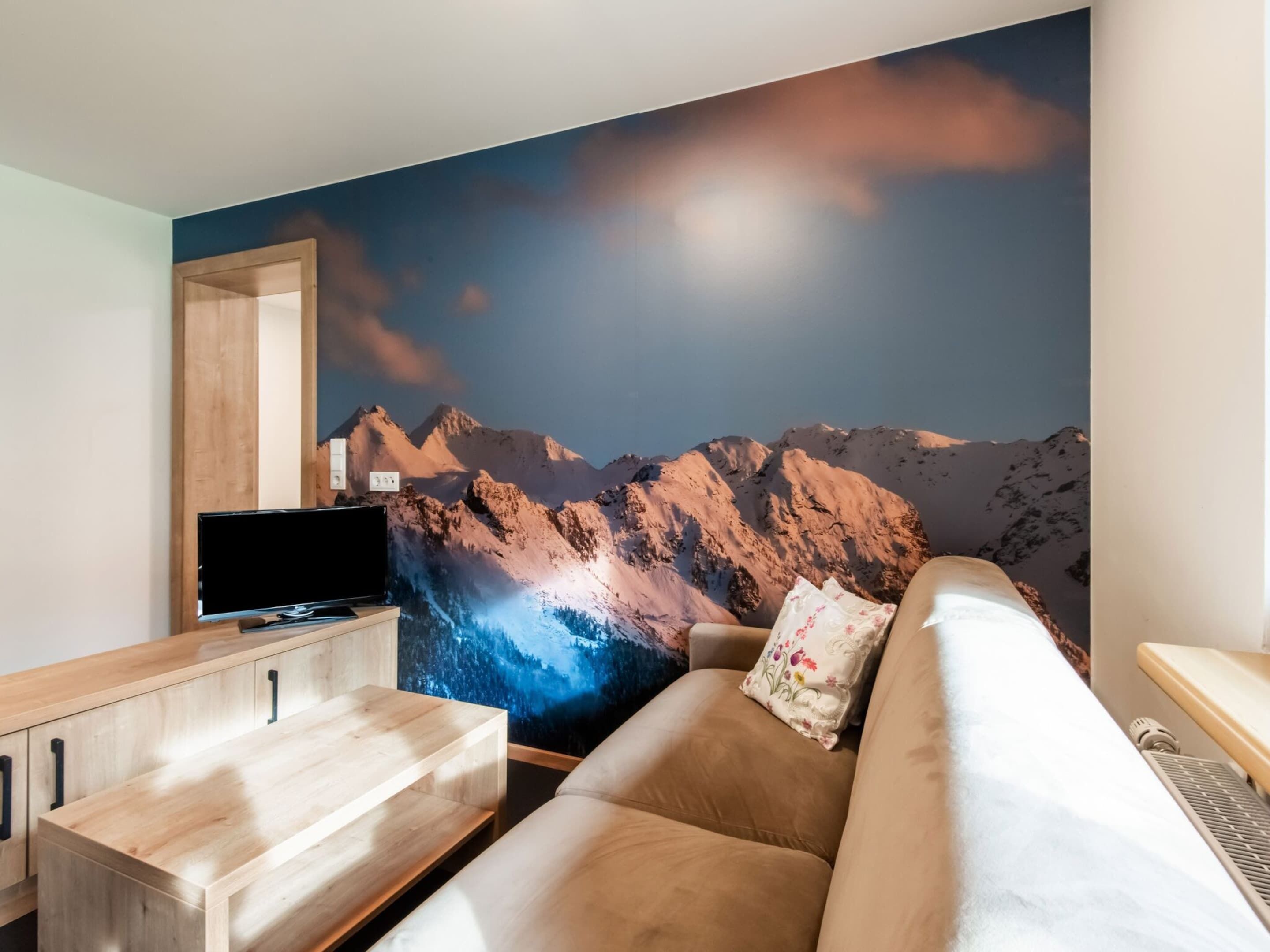 En moderne stue med et bjergmotiv, et tv-bord i træ og en beige sofa.