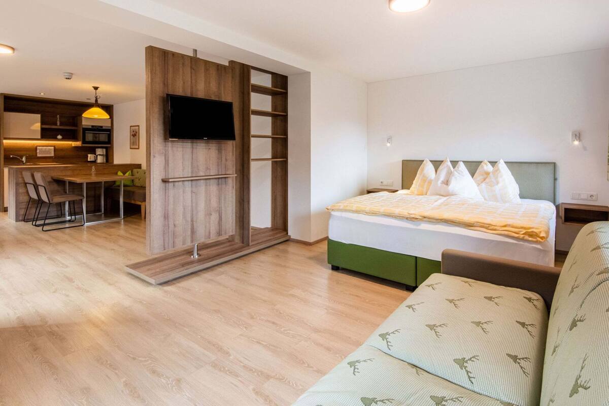 A modern hotel room with a bed, a sofa, a TV, a kitchenette, and a dining area.