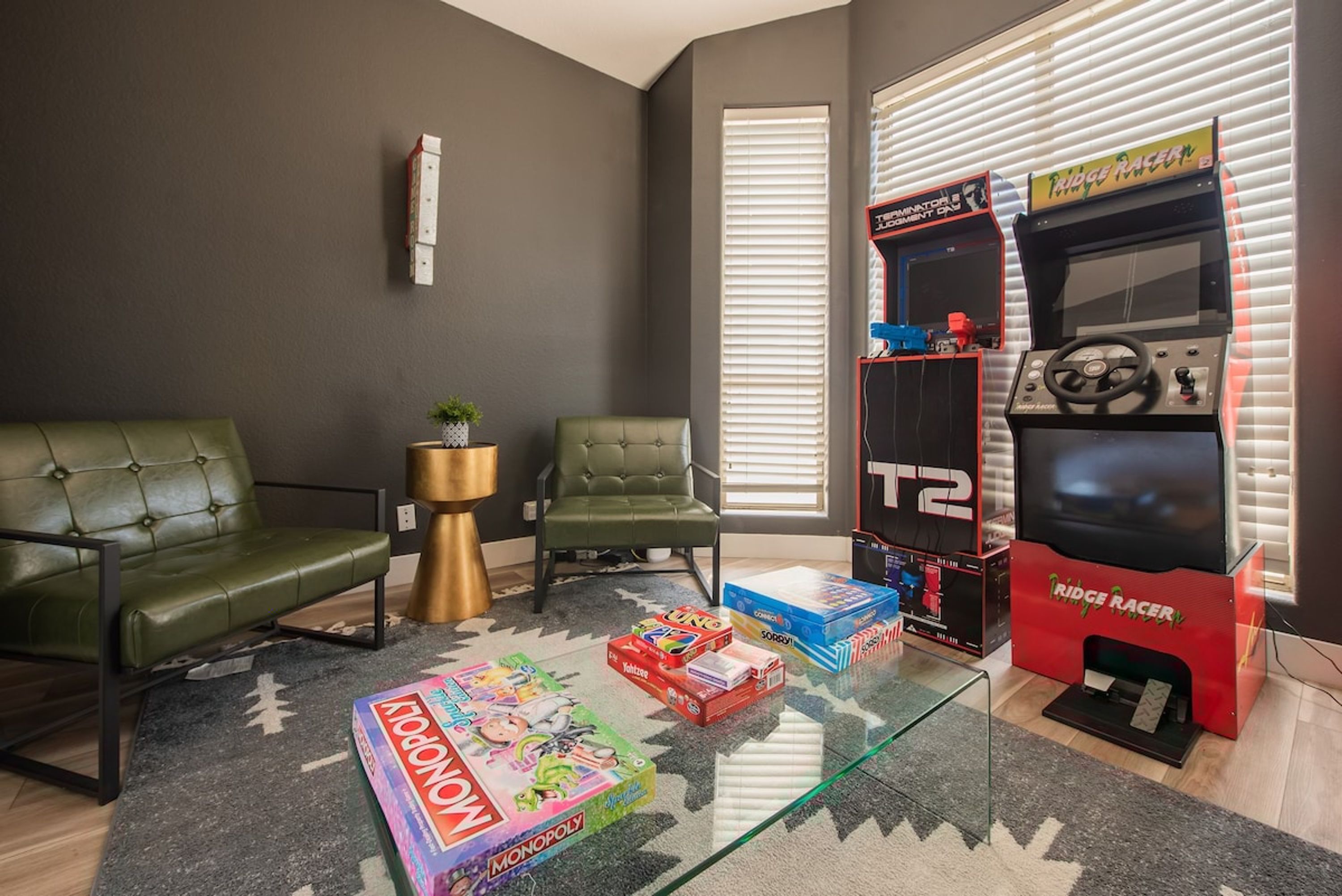 Ein Wohnzimmer mit einem grünen Sofa, zwei Sesseln, einem Glastisch mit Gesellschaftsspielen und einer roten Arcade-Maschine.