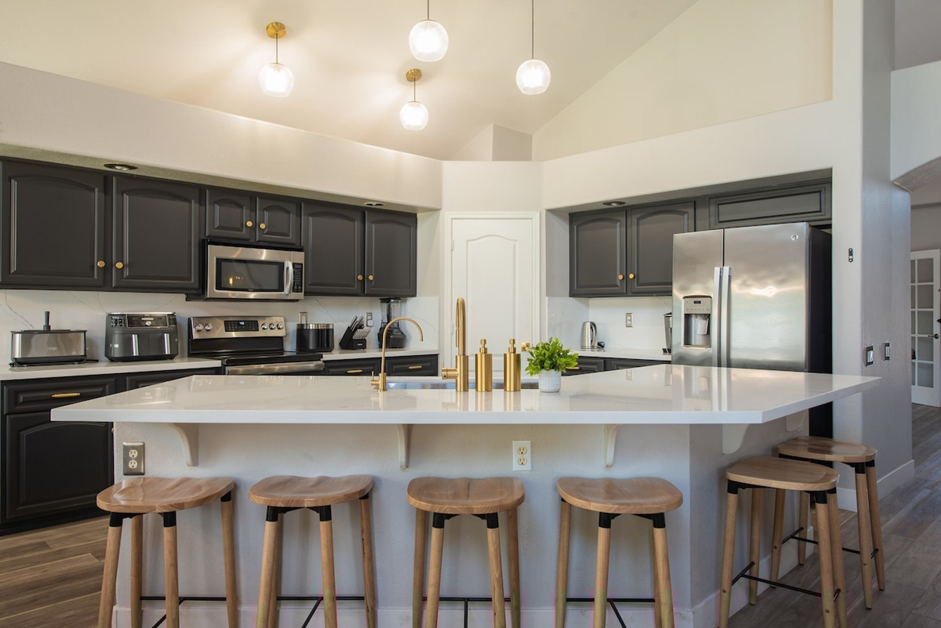 A modern kitchen with dark cabinets, stainless steel appliances, a large island with stools, and pendant lighting.
