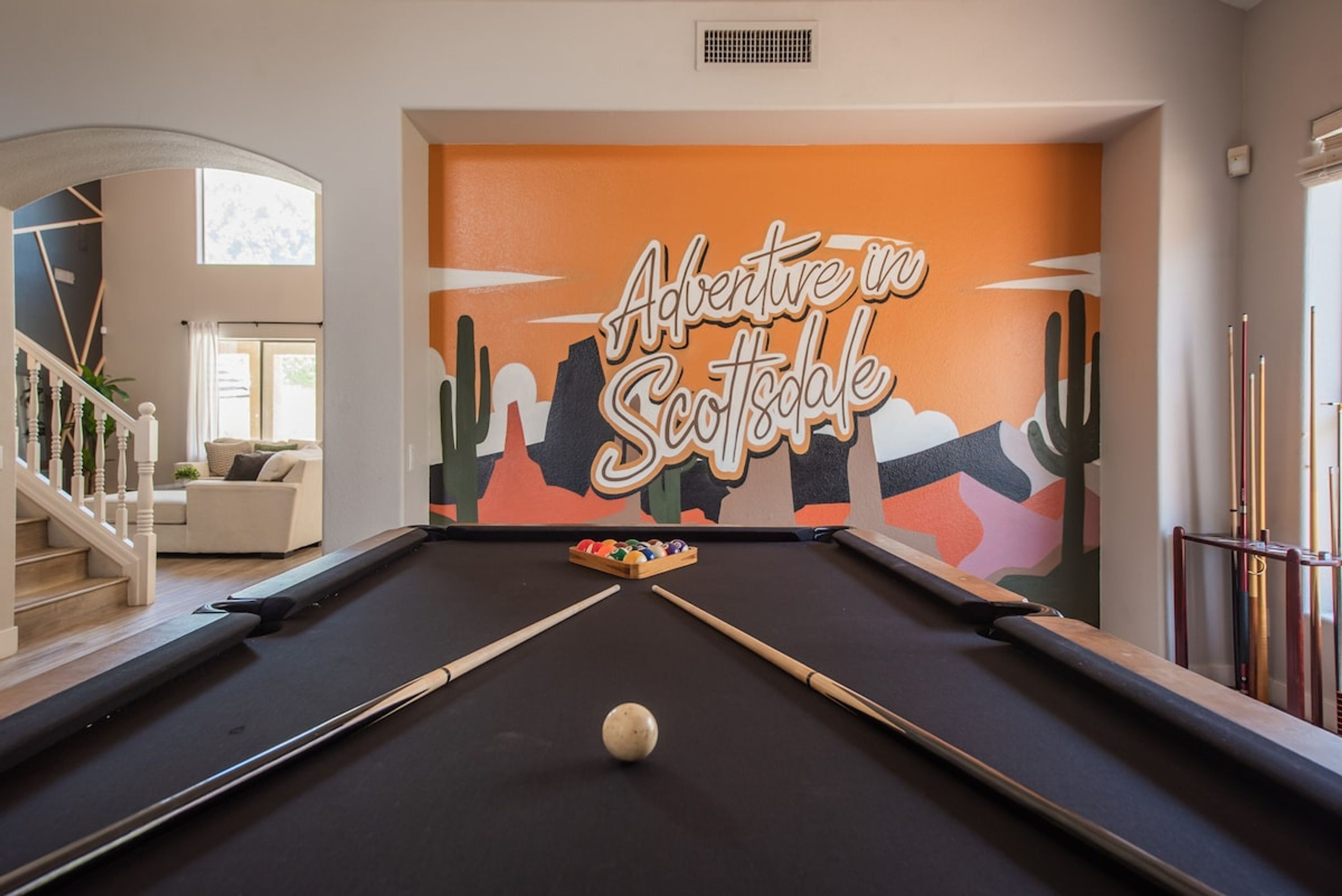 A pool table with a cue ball in the center, a pool table in a room with a mural reading "Adventure in Scottsdale" and a staircase leading to another room.