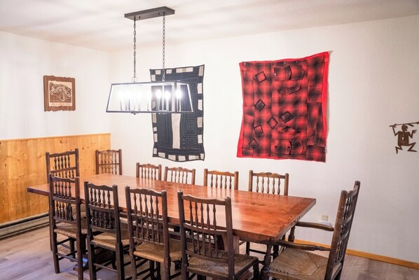 A dining area with a wooden table and chairs, a hanging light fixture, and a red and black patterned wall hanging.