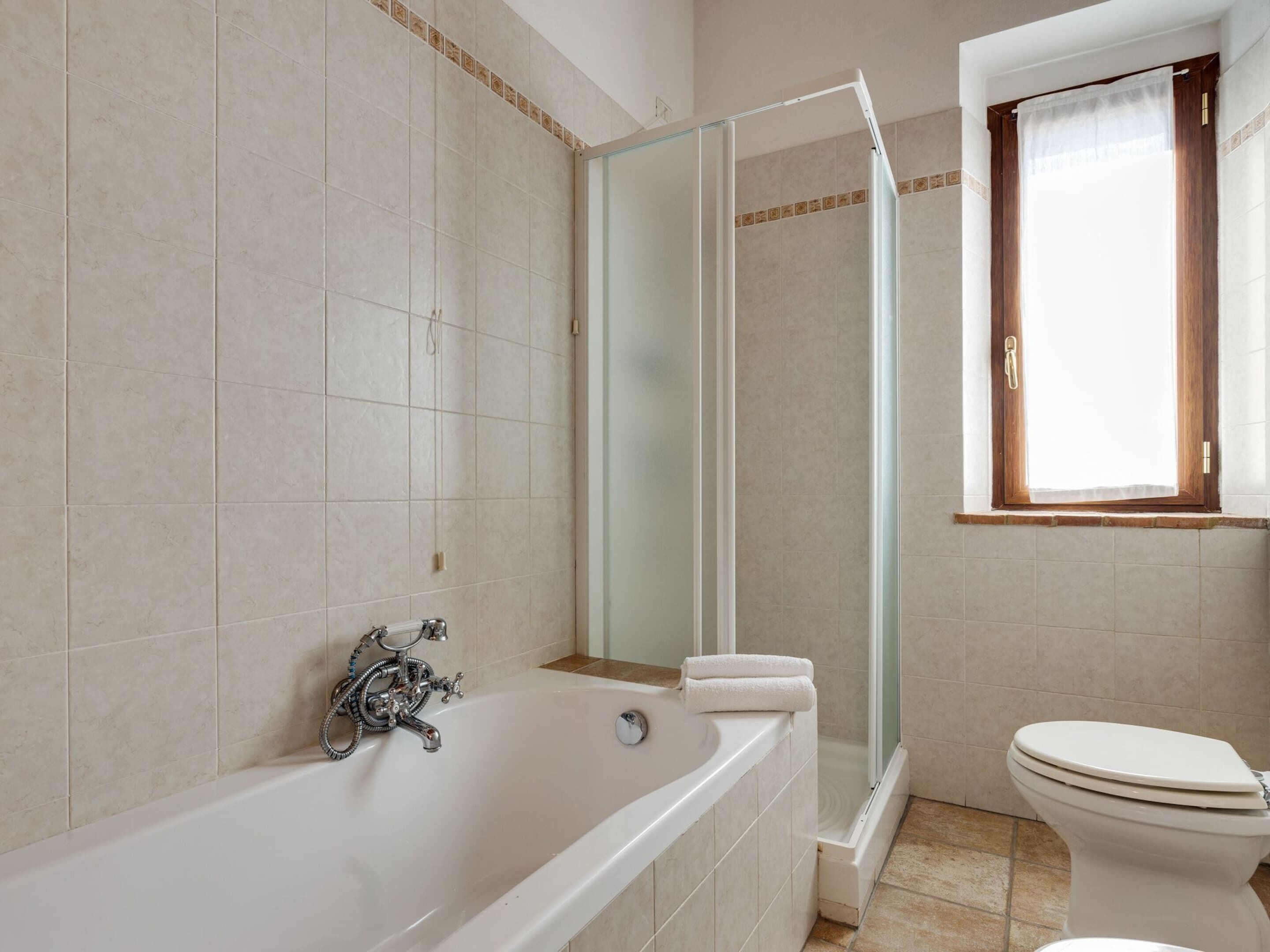 A bathroom with a bathtub, a glass-enclosed shower, a toilet, and a window with a curtain.