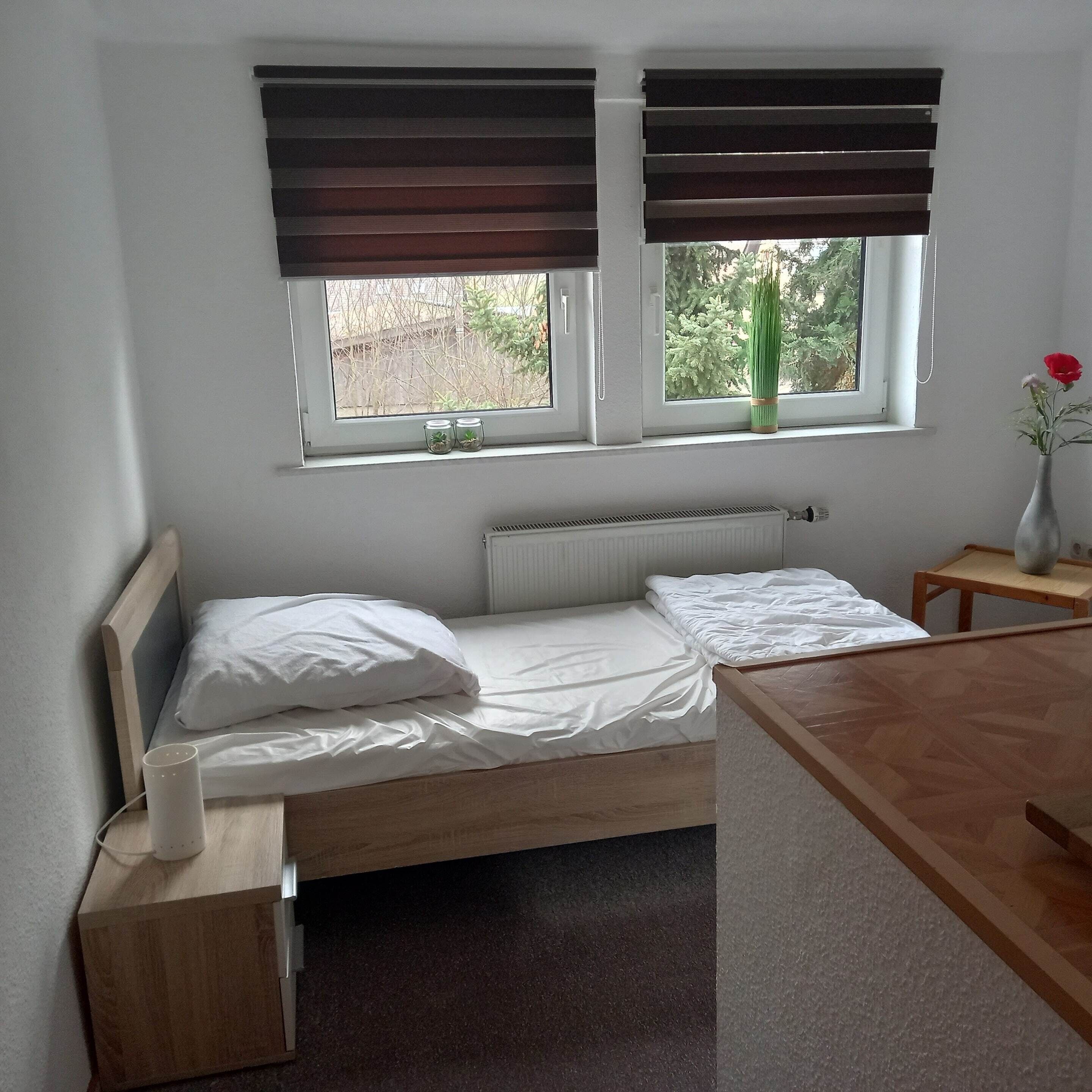 Una cama pequeña y bien hecha con sábanas blancas, cabecera de madera y mesita de noche con lámpara. Hay dos ventanas con persianas marrones, una de ellas con una planta pequeña en el alféizar.