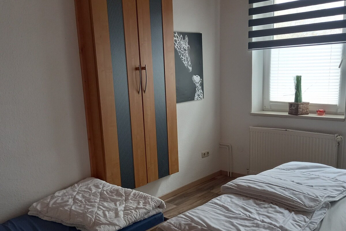 A bedroom with two single beds, a wardrobe, a window with blinds, and a radiator.