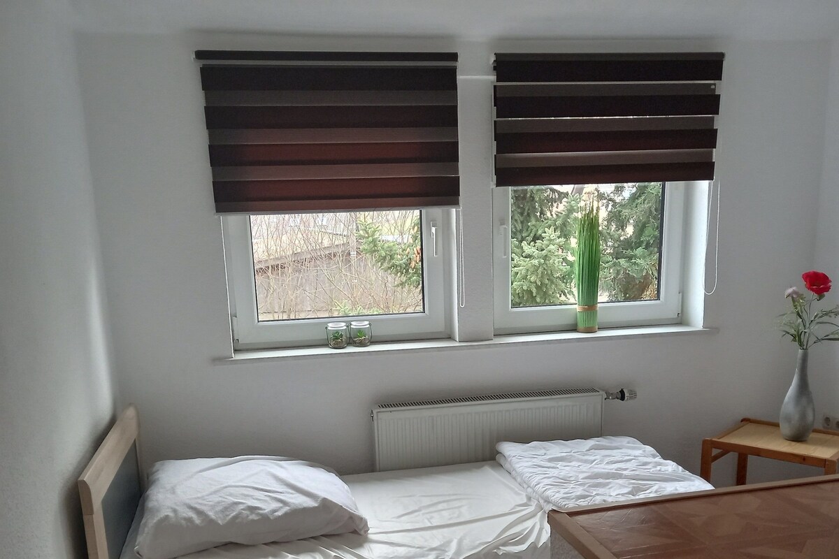A bedroom with a wooden bed, white bedding, a bedside table, a nightstand, a window with blinds, and a vase with flowers.
