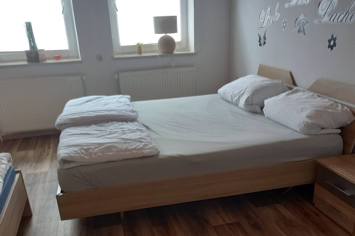 Ein ordentlich bezogenes Bett in einem Zimmer mit Holzdielen, einem Fenster mit Jalousien und einer Wand mit dekorativen Schmetterlingsstickern.
