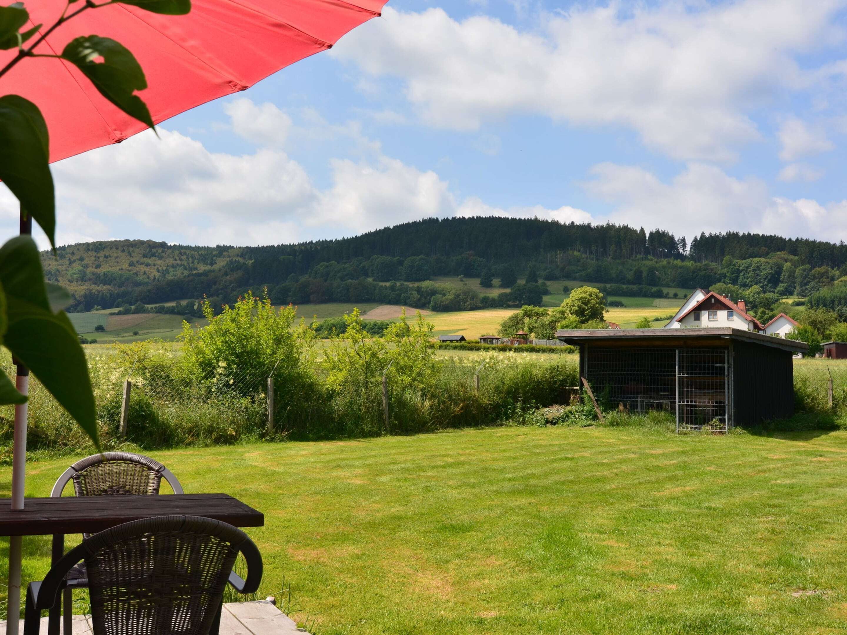 A grassy outdoor area with a table and chairs, a red umbrella, and a view of a hill and buildings in the background.