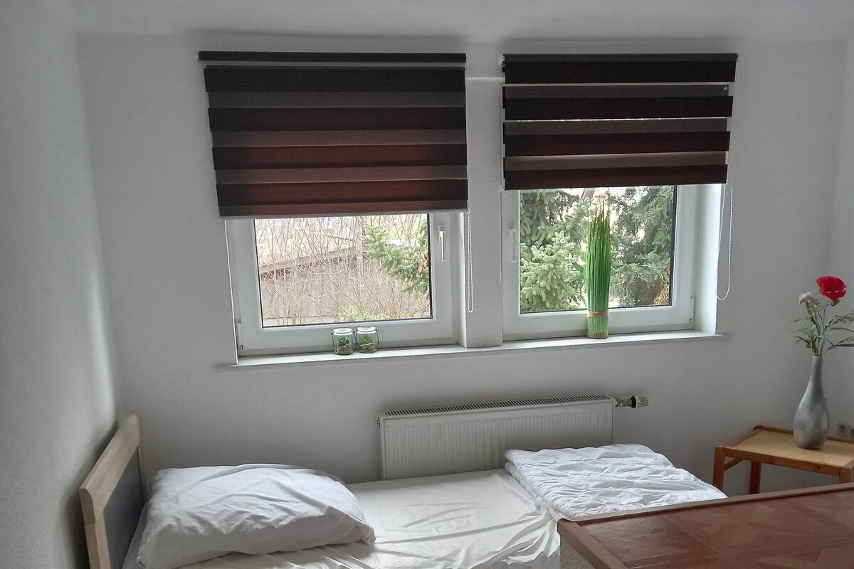 Una cama pequeña y bien hecha con sábanas blancas, cabecera de madera y mesita de noche con lámpara. Hay dos ventanas con persianas marrones, una de ellas con una planta pequeña en el alféizar.