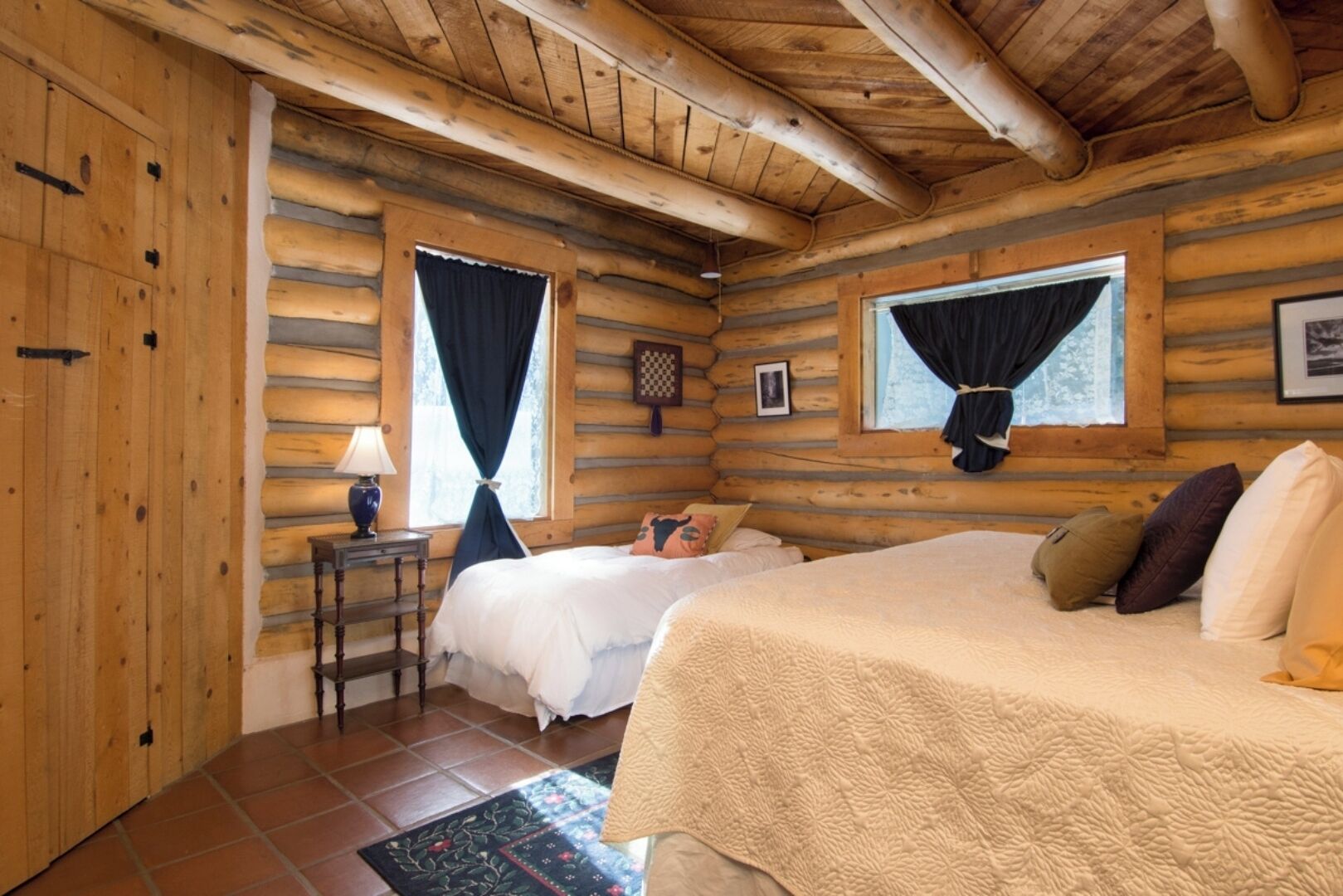 Une chambre aménagée dans un chalet en rondins, avec deux lits, une petite table et une fenêtre avec des rideaux.