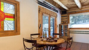 A cozy dining area with a wooden table and chairs, a stained glass window, and a view of a snowy landscape. - Denton Cabin - Warm Log Cabin, close to Mountain Stream and Ski Return Trail (Taos Ski Valley)