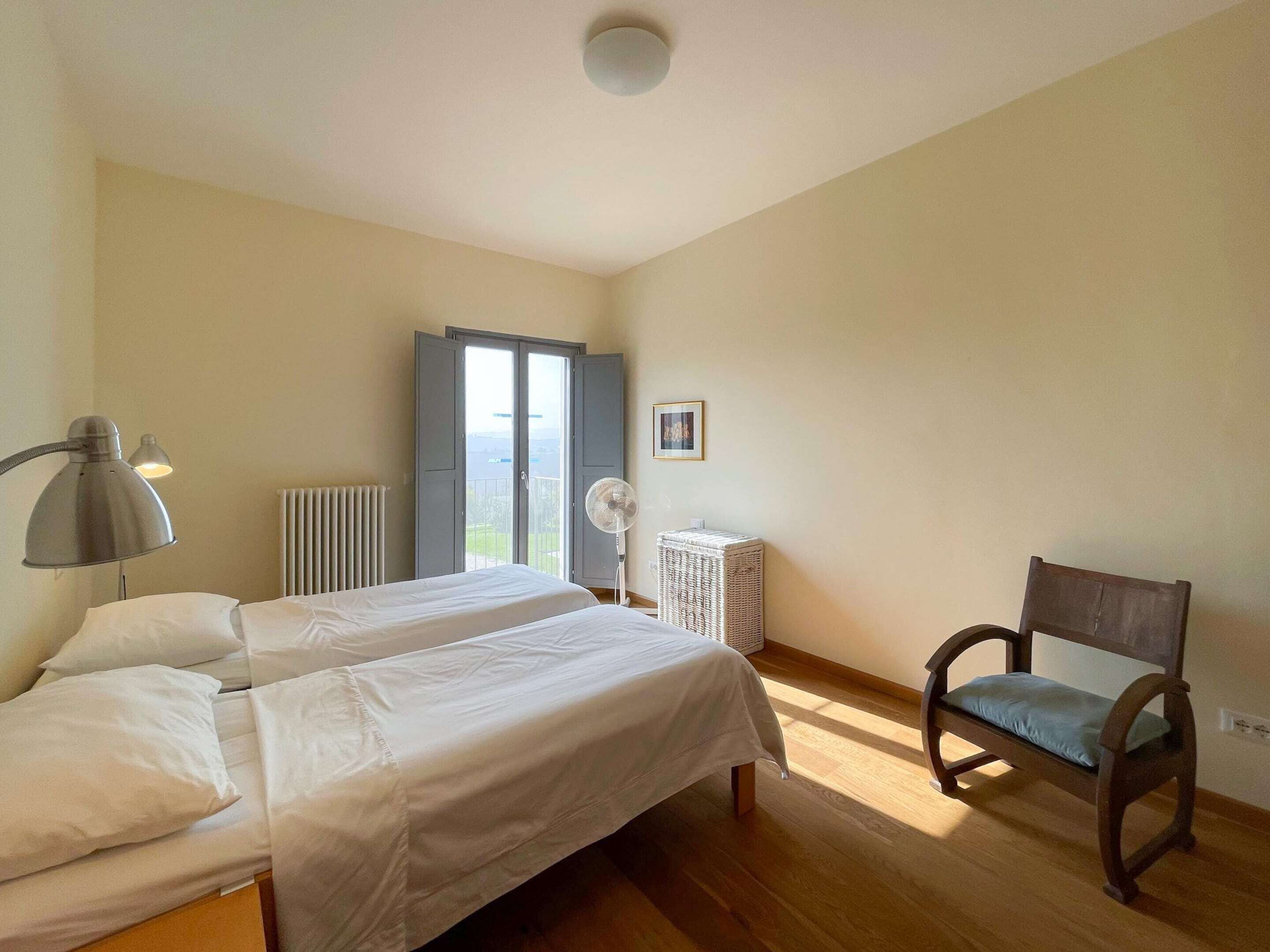 A bedroom with two beds, a chair, a radiator, and a window with curtains.
