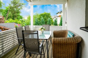 Eine überdachte Terrasse im Freien mit Tisch und Stühlen, ein Korbstuhl und Blick auf Grünflächen und Gebäude.