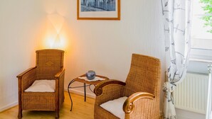 Quarto com duas cadeiras de vime, uma mesa pequena com um vaso, um quadro na parede e uma janela com cortina.