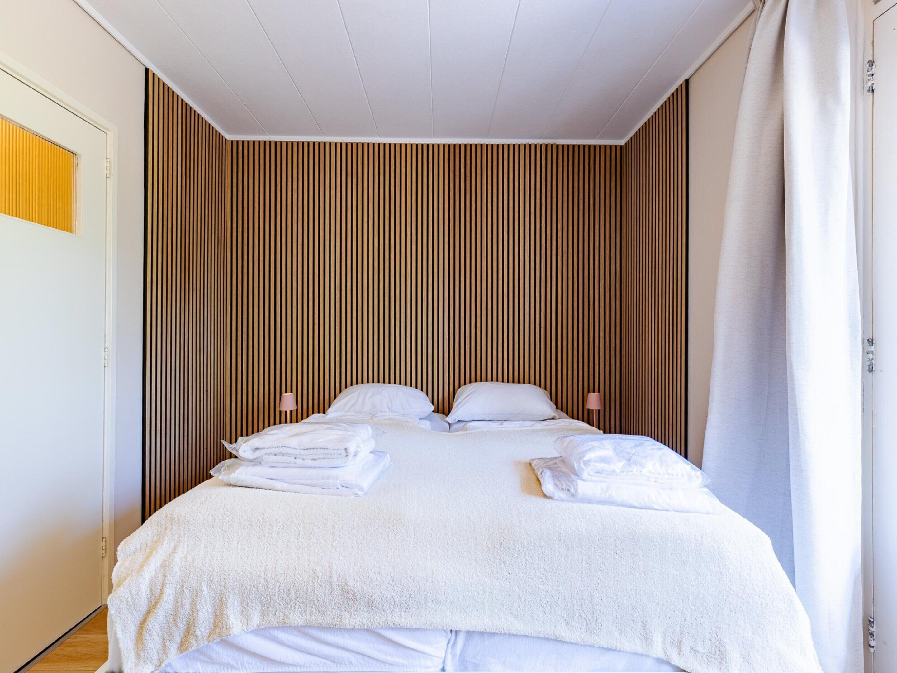 A neatly made bed with white linens and towels, a wooden slat wall, and a white ceiling.