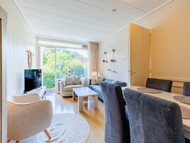 A modern living room with a TV, sofas, a dining table, and a view of greenery outside.