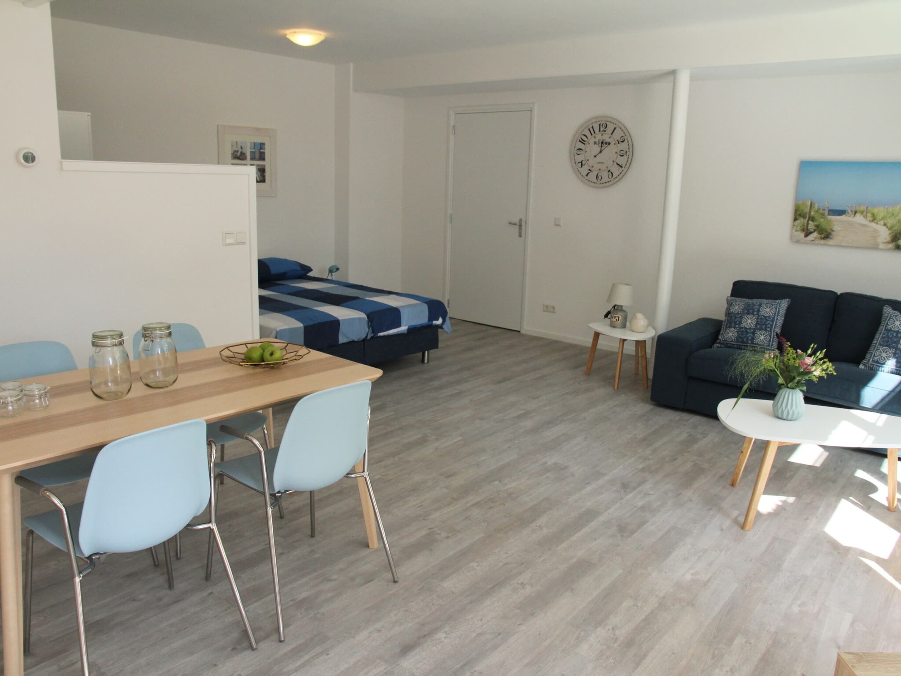 Un salon moderne comprenant un canapé, une table à manger avec des chaises, un lit avec une couvre-lit à rayures bleues et blanches, et une horloge murale.