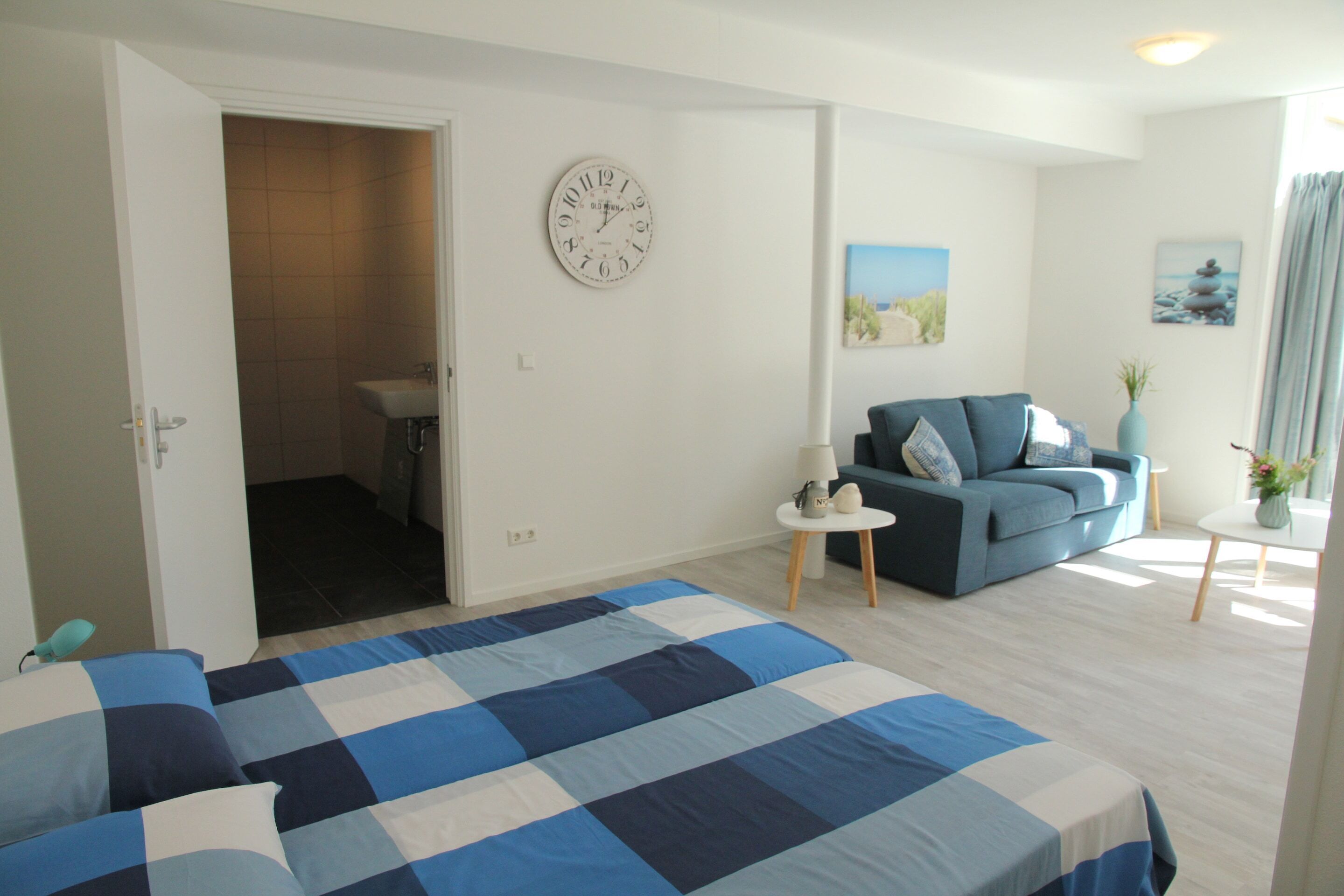 A bedroom with a checkered bedspread, a blue sofa, a round wall clock, and a small side table with a lamp.