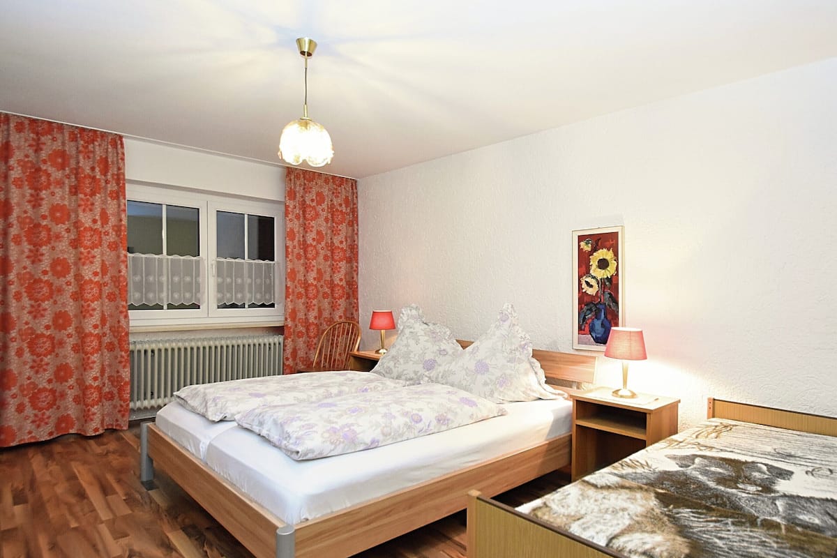 Ein Schlafzimmer mit einem hölzernen Bett, einem Nachttisch mit Lampe, einem Fenster mit Vorhängen und einem Bild an der Wand.