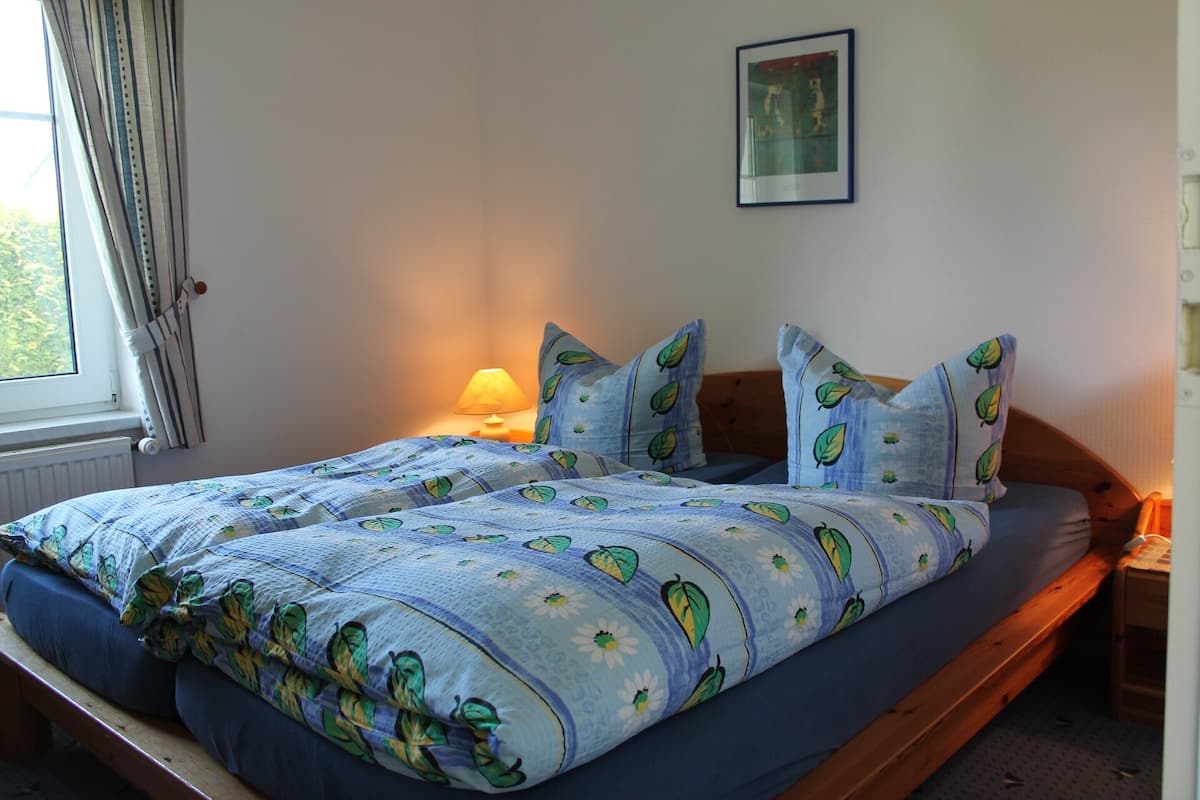 A double bed with a leaf-patterned duvet, a bedside table with a lamp, and a window with curtains.